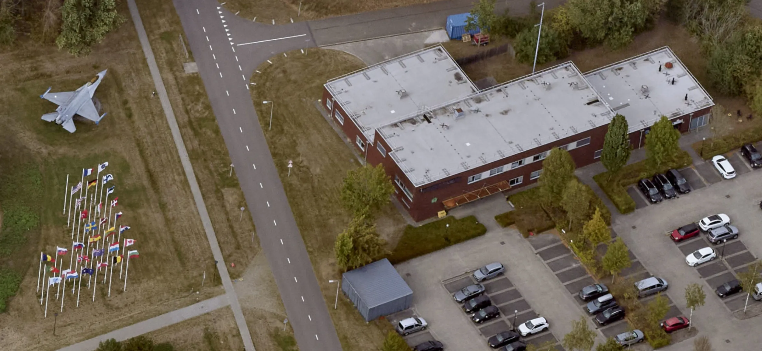 Aerial view of a building at Vliegbasis Eindhoven with a fighter aircraft on display and a row of flags lining the access road.