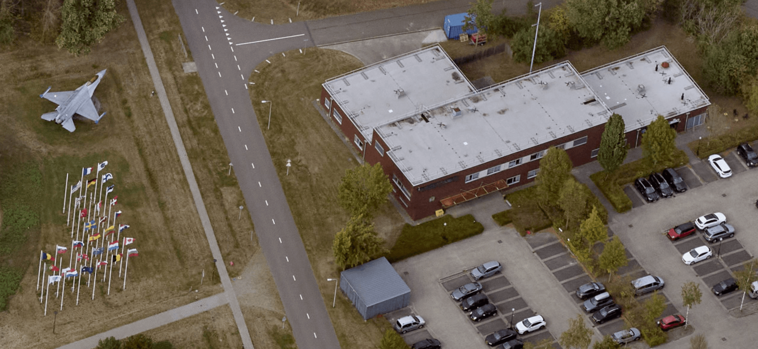 Aerial view of a building at Vliegbasis Eindhoven with a fighter aircraft on display and a row of flags lining the access road.